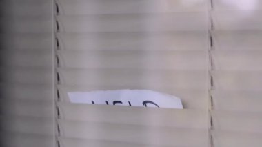 Adam panjurların arasından bir kağıt parçasına yazılmış yardım çağrısını gösteriyor. Panjurlarla kapatılmış bir pencerede yardımla ilgili bir yazı olan bir kağıt parçası belirir