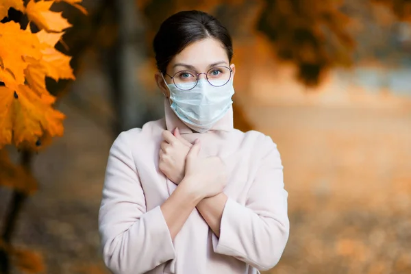Yüz maskesi takmış genç bir kadın ellerini göğsünün üzerine koyup yaklaşan soğuk algınlığının ve hastalığın sembolü olarak ceketinin yakasını sıkıca kapatıyor. — Stok fotoğraf