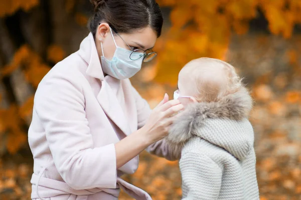 Femme dans un masque facial médical ajuste le masque médical à la petite fille, étant dans la cour à l'automne — Photo