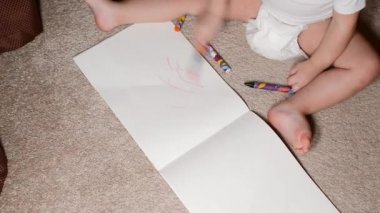Çocuk halıya oturur ve bir albümde pastel boya çizer, yüzü yoktur.