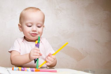 Gülümseyen bir bebek elinde tuttuğu kalemlere ve pastel boyalara bakar.