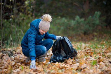 Genç bir kadın sonbahar ormanında çöp topluyor.