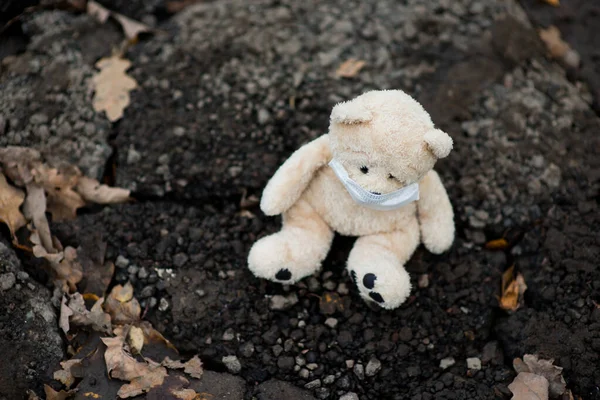 A teddy bear in a medical mask sits on dirty ground — Stock Photo, Image