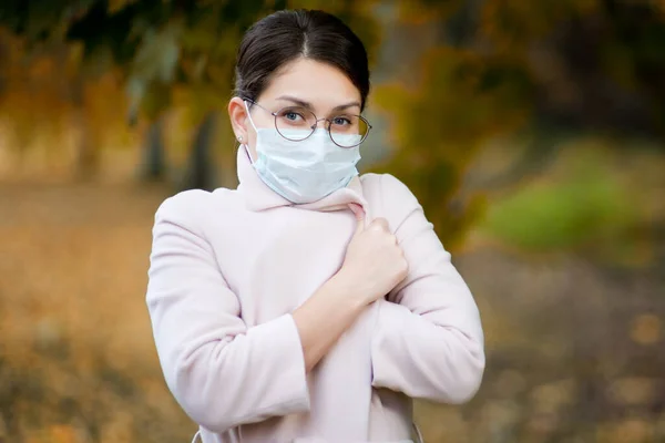 Mujer joven con una máscara médica envuelta en un abrigo ligero en un día fresco de otoño. De cerca. —  Fotos de Stock