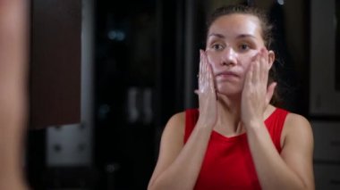 Genç bir kadın banyoda aynanın önünde yüzünü yıkıyor. Kapat.