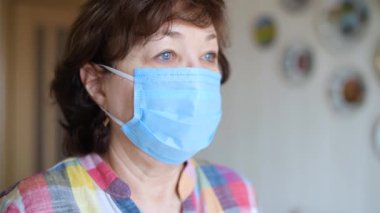 Koruyucu tıbbi maskeli yaşlı bir kadın pencerenin önünde duran ışığa bakıyor. Kamera yavaşça yana kayar..