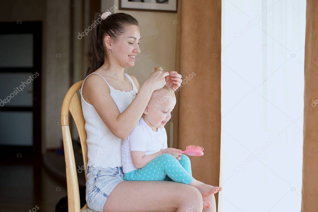 A young mother is having a great time with her daughter. Mom makes a ponytail on the head of a little girl who sits on her mother s lap near the window of the house
