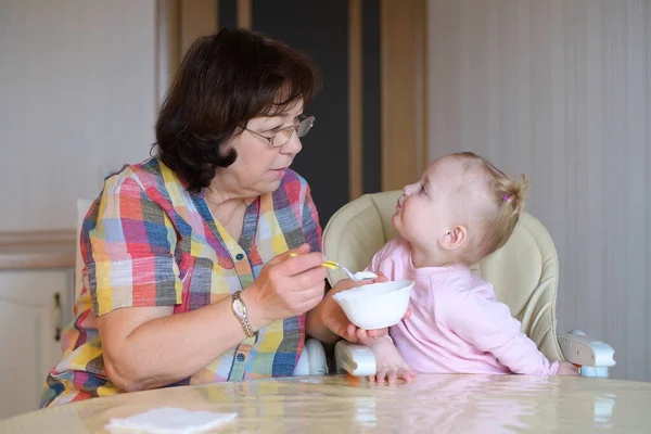 General view of the grandmother and granddaughter in the process of feeding yogurt. Nanny and little child eat food at the table in the kitchen