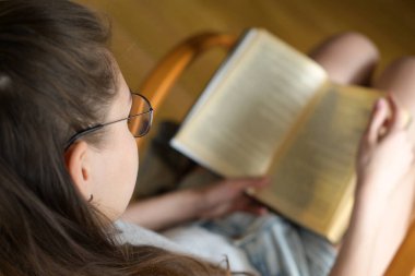 Gözlüklü bir kız gün ışığında kitap okuyor. Gözlüklere odaklan. Üst görünüm