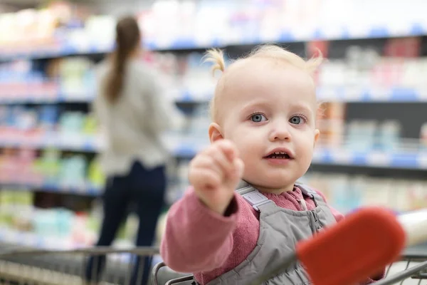 little girl sitting in a grocery cart and waiting for mom to choose groceries in a supermarket