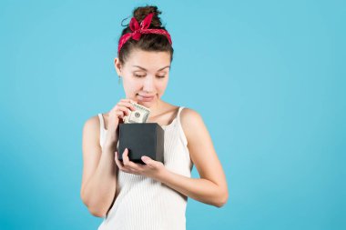 Genç bir kadın mavi arka plandaki bir tasarruf kutusuna dolar koyar ya da çeker.