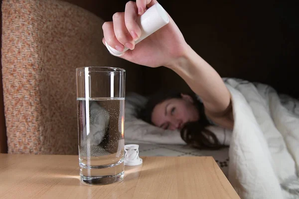 一个年轻的女人很难把含有止疼药或宿醉药的可溶性药丸放在放在放在床头柜的一杯水里 选择性地聚焦在玻璃上 — 图库照片
