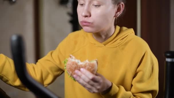 Dziewczyna bezczynnie trenuje na maszynie sportowej i żuje hamburgera — Wideo stockowe