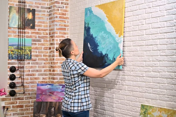 A girl hangs a picture on the wall in her creative studio, then moves away and admires the creation. the artist in the studio looks at the picture