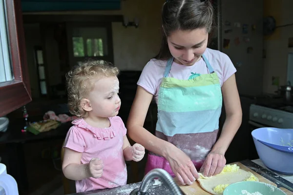 Küçük bir kız, yemek hazırlayan annesinin yanında, mutfak lavabosunda unla oynuyor. Neşeli ev rutini, pencereden bakmak.