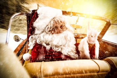 Noel Baba, güneşli bir kış gününde Noel hediyelerini teslim etmek için arabaya biniyor. Kırmızı arkaplanda Noel süslemesi.