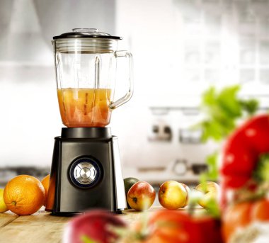 Masanın üzerinde biraz meyve ve sebze ve bulanık mutfak arkaplanı olan bir blender var..
