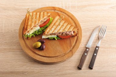 Tahta tepsiye ton balığı dolgusu yla kızarmış beyaz ekmekkulübü sandviçi. Sandviç ikiye bölünür. Yakınlarda birkaç zeytin var. Açık ahşap bir yüzey çatal üzerinde. Close.