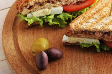 Beyaz tost ekmeğinden ton balığı ile sandviç. Ton balığı, yumurta, domates, yeşil salata. Sandviç tahta yuvarlak tepside. Yakınlarda birkaç zeytin var. Yukarıdan görüntüleyin. Makro fotoğrafçılık.