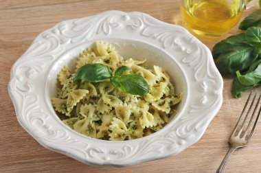 Beyaz bir tabakta pesto soslu makarna farfalle. Yemek taze fesleğen yaprağı ile dekore edilmiştir. Tabağın yanında bir çatal, bir sürahi zeytinyağı ve bir demet taze fesleğen var. Close.