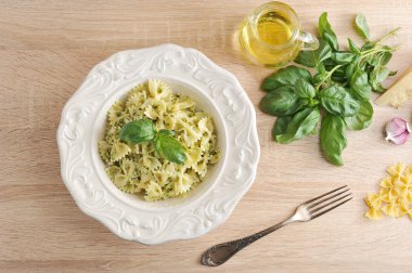 Beyaz bir tabakta pesto soslu makarna farfalle. Sonraki çatal, bir sürahi zeytinyağı, bir demet taze fesleğen, badem, parmesan peyniri, sarımsak ve makarna. Close. Açık ahşap arka plan.
