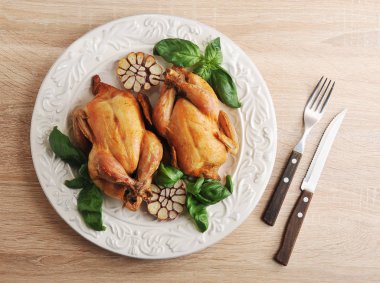 Izgarasında iki tavuk pişirilmiş. Sarımsak ve taze fesleğen yeşillikleri yarım beyaz bir tabak üzerinde. Yakınlarda çatal bıçak takımı var. Ahşap arka plan. Close. Yukarıdan görüntüle. 