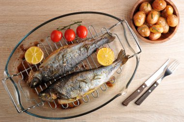 İki nehir alabalığı büyük bir tabakta ızgara. Yemek pişirirken limon ve çeri domates kullanılmıştır. Bir sonraki çatal bıçak takımı ve mini patatesli bir kase. Yukarıdan görüntüleyin. Close. 