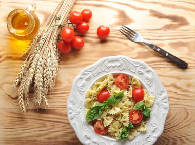 Pesto soslu makarna farfalle ve çeri domates. Makarna taze fesleğen yaprakları ile dekore edilmiştir. Arka planda, buğday kulakları, zeytinyağı, domates ve çatal. Açık ahşap arka plan. Close.