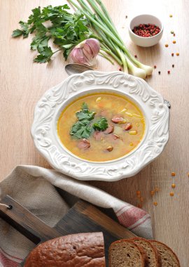 Füme sosis ve maydanoz lu bezelye çorbası. Tabağın yanında bir dizi baharat var: yeşil soğan, sarımsak, maydanoz, dolmalık biber. Doku peçetesinin kenarı görünür. Close.