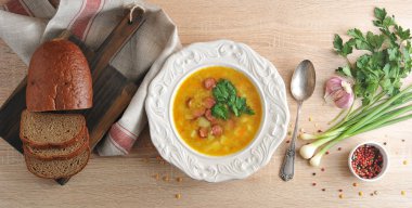 Beyaz tabakta füme etli bezelye çorbası. Ekmek ahşap bir tahta ve bez peçete üzerinde dilimlenmiş. Tabağın yanında, bir kaşık ve baharatlar: sarımsak, maydanoz, yeşil soğan ve biber. Yukarıdan görüntüle.