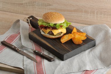 Tahta tahtada pastırmalı sulu çizburger. Sonraki patates dilimleri rustik. Çerçevede bir doku peçete ve çatal bıçak takımı var.