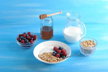 Sağlıklı bir kahvaltı servis: müsli, ahududu ve yaban mersini, bal, cam sürahi yoğurt. Mavi ahşap arka plan. Close.