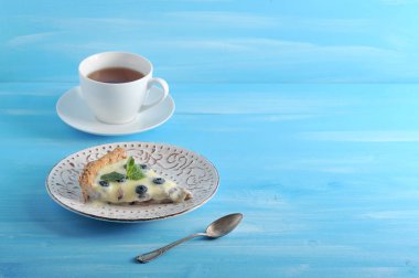 Açık bir kum pasta bir kısmı - desenleri ile bir tabak üzerinde beyaz çikolata ve yaban mersini ile bir tart. Mavi ahşap arka plan. Çay ve bir çay kaşığı ile sonraki fincan.