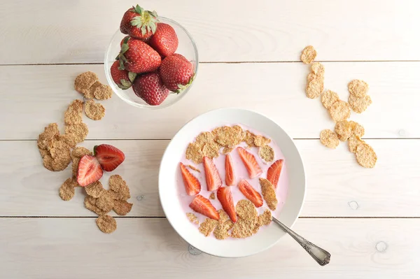 Dark corn flakes with milk and strawberries. Delicious and healthy breakfast. In a plate with flakes spoon.  Next to the plate with strawberries. Light wooden background. View from above.
