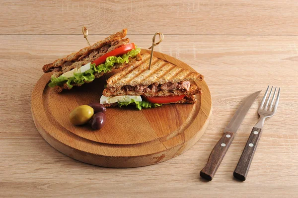 Tahta tepside, ton balığı dolgulu kızarmış beyaz ekmekli bir sandviç. Sandviç ikiye bölünür. Yakınlarda birkaç zeytin var. Açık ahşap bir yüzey çatal üzerinde. Close.