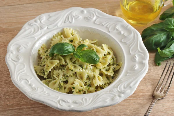 Beyaz bir tabakta pesto soslu makarna farfalle. Yemek taze fesleğen yaprağı ile dekore edilmiştir. Tabağın yanında bir çatal, bir sürahi zeytinyağı ve bir demet taze fesleğen var. Close.