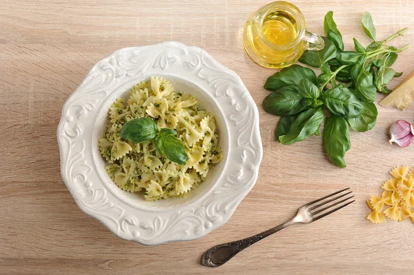 Beyaz bir tabakta pesto soslu makarna farfalle. Sonraki çatal, bir sürahi zeytinyağı, bir demet taze fesleğen, badem, parmesan peyniri, sarımsak ve makarna. Close. Açık ahşap arka plan.