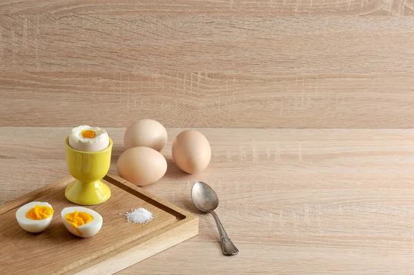 Ahşap bir tepsiüzerinde sert haşlanmış yumurta, yumuşak haşlanmış yumurta ve tuz iki yarısı vardır. Bir çay kaşığı ve yumurta kabuğunda. Açık ahşap arka plan. Metin için bir yer var.