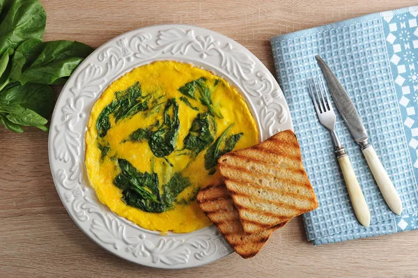 Omelette with spinach on a plate and two toast. Next cloth napkin and cutlery. Behind the plate is a bunch of fresh spinach. Light wooden background. View from above.