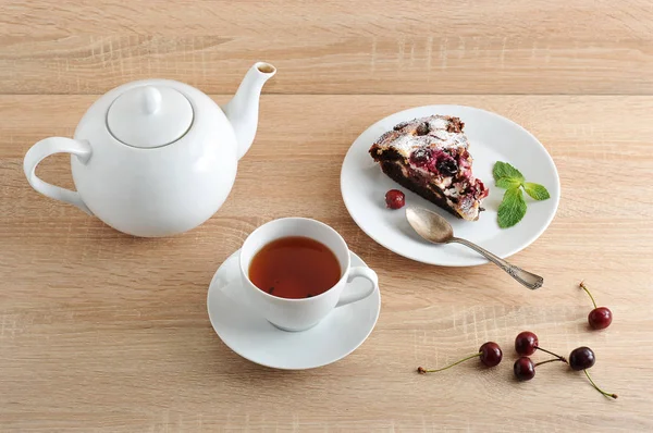 Bir tabakta çikolatalı kek parçası. Pasta nane yaprağı ile dekore edilmiştir. Bir sonraki fincan çay ve çaydanlık. Taze kirazlar masada. Bir kişiye hizmet ediyor. Açık ahşap arka plan.
