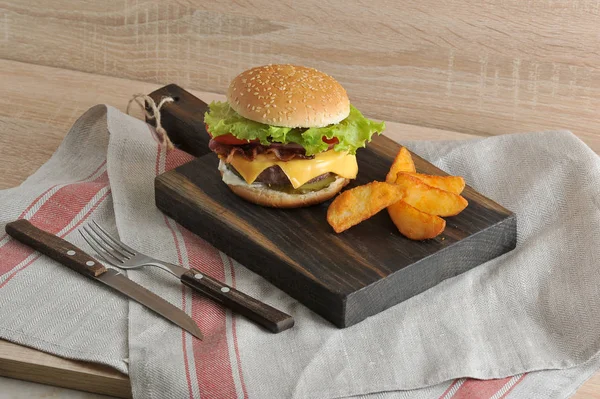 Tahta tahtada pastırmalı sulu çizburger. Sonraki patates dilimleri rustik. Çerçevede bir doku peçete ve çatal bıçak takımı var.