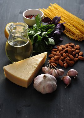 Pesto soslu makarna için malzemeler. Çerçevede yeşil ve mor fesleğen, badem, parmesan peyniri, sarımsak, baharat, zeytinyağı ve Fusilli lunghi makarna. Karanlık arka plan. Close. Dikey çekim.