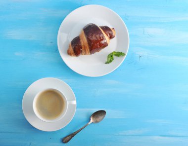 Çikolatalı kruvasan ve bir fincan kahve kahvaltı veya atıştırmalık için mükemmel bir kombinasyondur. Taze nane bir kruvasan yaprağı ile bir tabak üzerinde. Bir sonraki çay kaşığı. Mavi ahşap arka plan. Close.