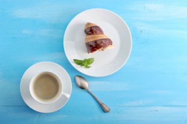 Bir fincan sade kahve ve yarım çikolatalı kruvasan. Taze nane bir kruvasan yaprağı ile bir tabak üzerinde. Bir sonraki çay kaşığı. Mavi ahşap arka plan. Close.