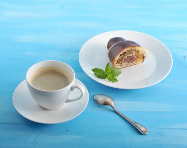 Yemyeşil ve havadar çikolatalı kruvasan ve bir fincan sade kahve. Tabakta, yarım kruvasan ve nane yaprağı. Bir sonraki çay kaşığı. Mavi ahşap arka plan. Close. 