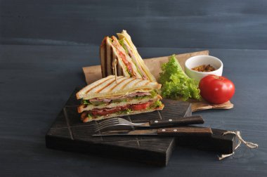 Tahta tahtada yarım sopalı sandviç. Sandviçin diğer yarısı kraft kağıdın yanında. Çerçeve çatal, hardal, marul, domates. Karanlık arka plan. Close. 