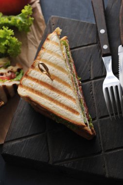 Tahta bir tahta üzerinde üçgen şeklinde bir sandviç yarısı. Sandviç marul yaprağı ile dekore edilmiştir. Yakınlarda çatal bıçak takımı var. Koyu ahşap arka plan. Close. Yukarıdan görüntüle.