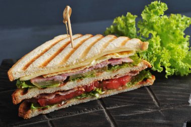 Tahta bir tahta üzerinde üçgen şeklinde bir sandviç yarısı. Sandviç marul yaprağı ile dekore edilmiştir. Dolgu jambon, peynir, domates, pastırma, sos ve yetenekli bir yumurta oluşur. Close.