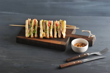 Ahşap bir tahta üzerinde, bir sandviç çapraz dört parçaya kesilir. Parçalar ahşap bir şiş üzerine sabitlenir. Bir sonraki çatal bıçak takımı ve bir fincan hardal. Koyu ahşap arka plan. Close.