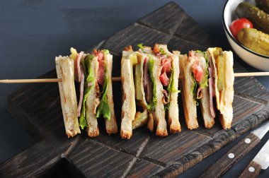 Tahta bir tahtanın üzerinde dört parçalık bir sandviç, tahta bir şiş üzerine sabitlenmiş. Sandviçin dolgusu pastırma, peynir ve jambondan oluşur. Koyu ahşap arka plan. Close.